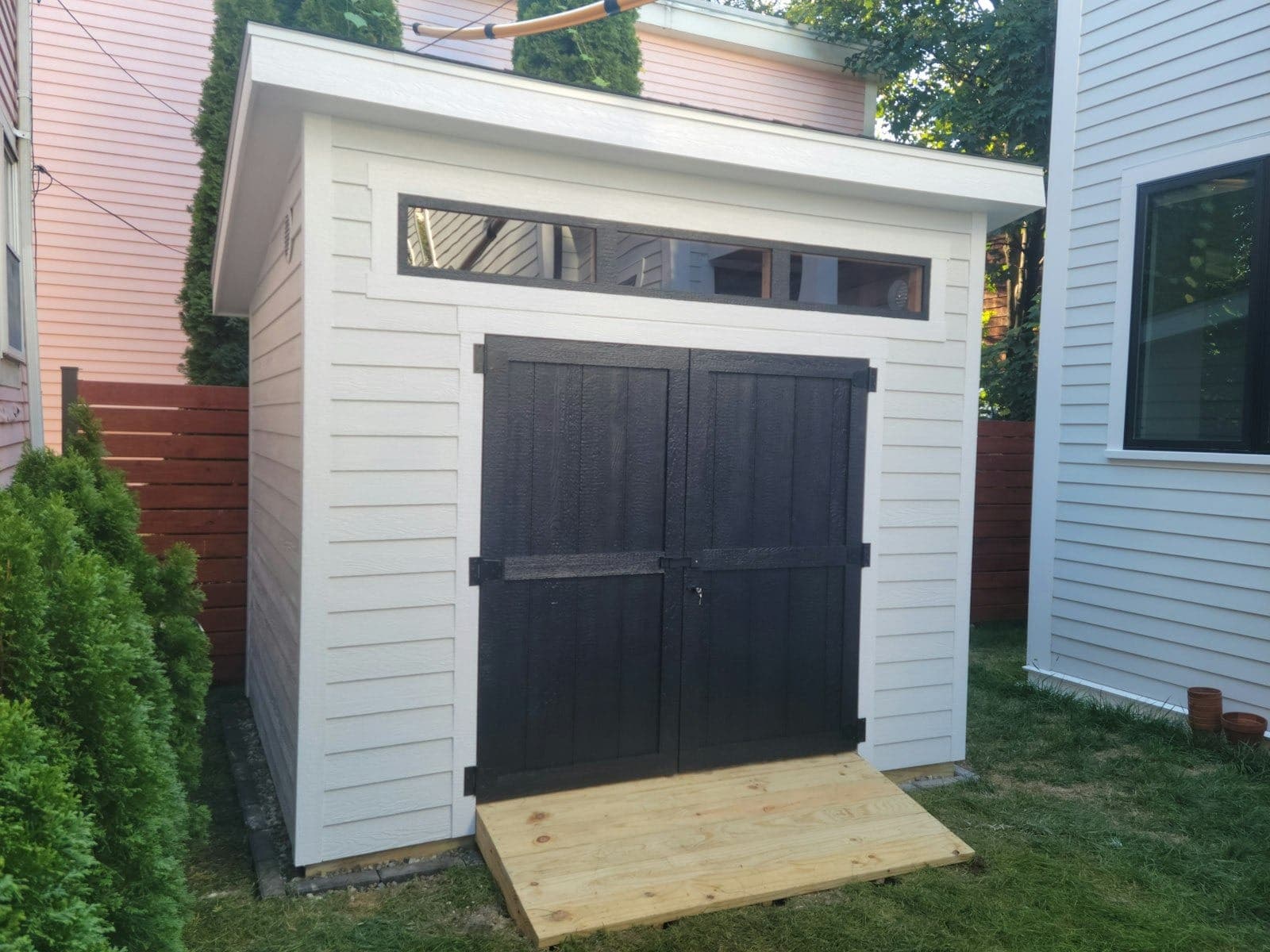A white lap-sided shed with black doors tucked between two houses in a narrow city yard
