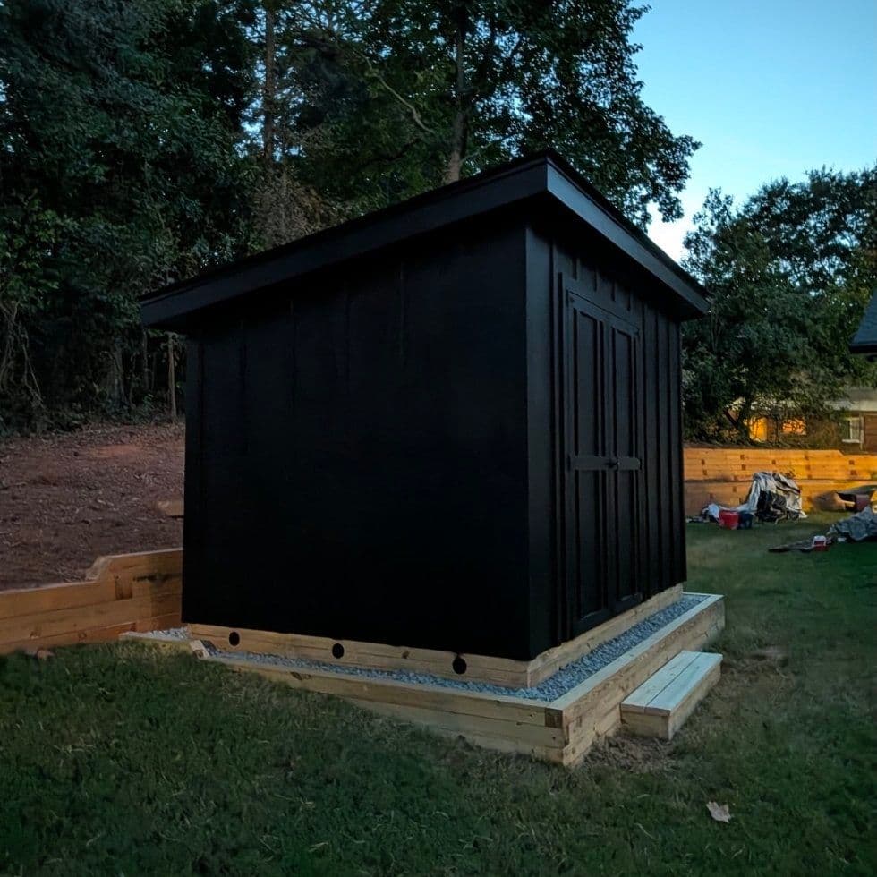 A black board-and-batten shed with a single-slope roof and double doors, on a stone pad with a timber perimeter, beside a raised garden bed