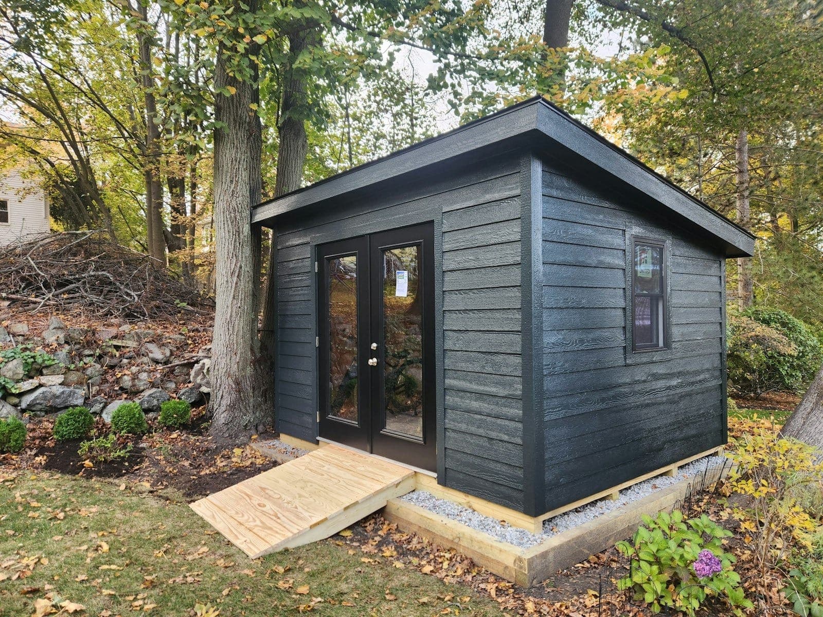 A black lap-sided shed with French doors and a wood ramp under autumn trees