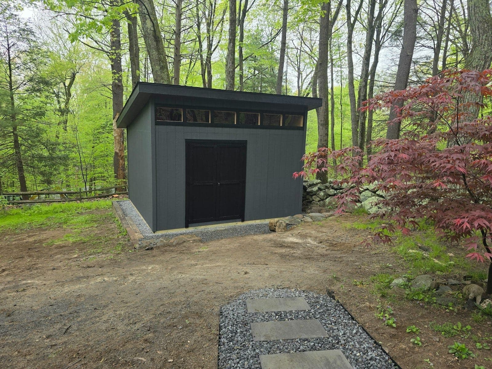 A dark modern shed with a flat roof and black double doors, built among tall trees beside a stone wall