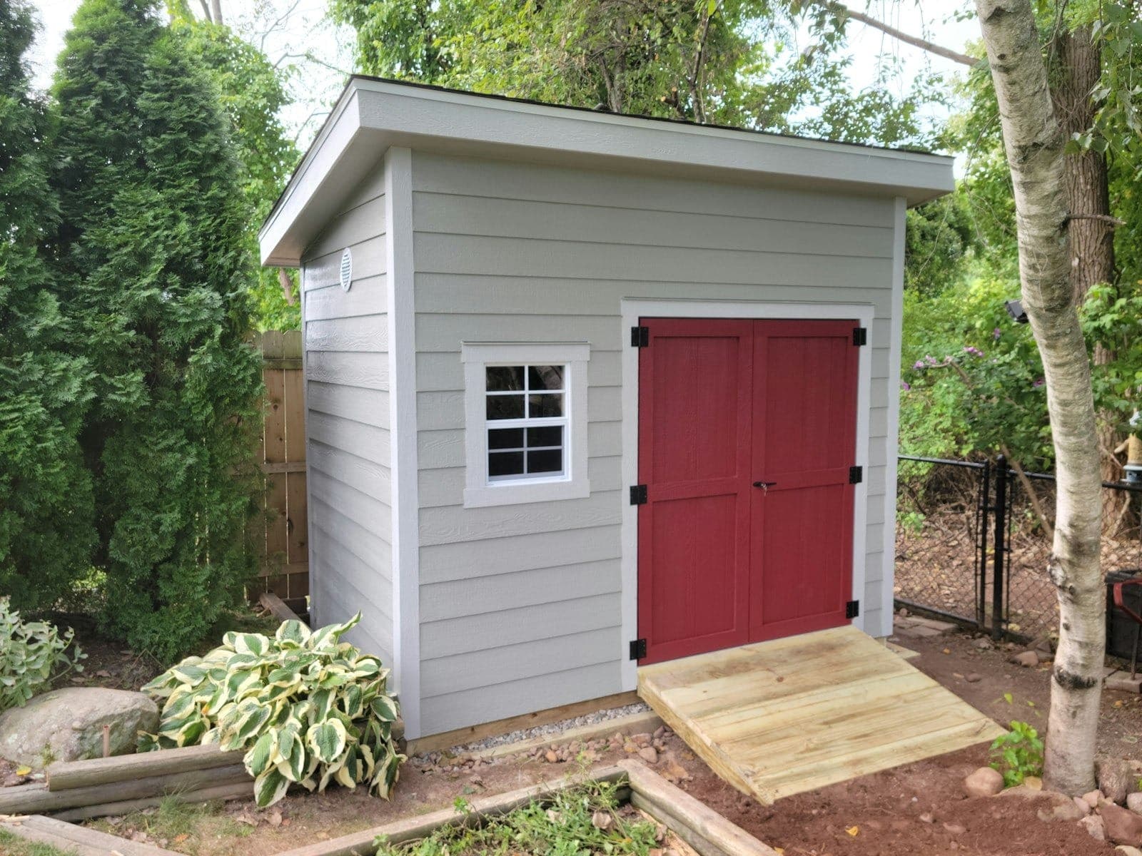 A gray lap-sided shed with a red double door and a wood ramp, beside a row of evergreens