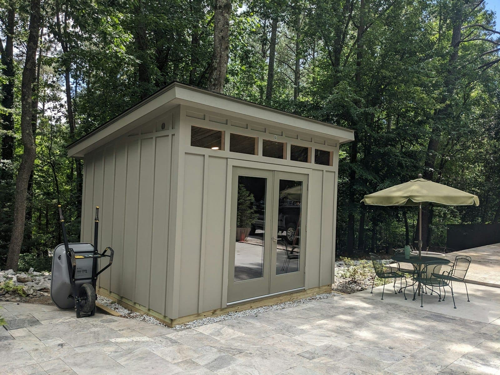 A taupe board-and-batten modern shed with French doors and a transom window row, beside a stone patio