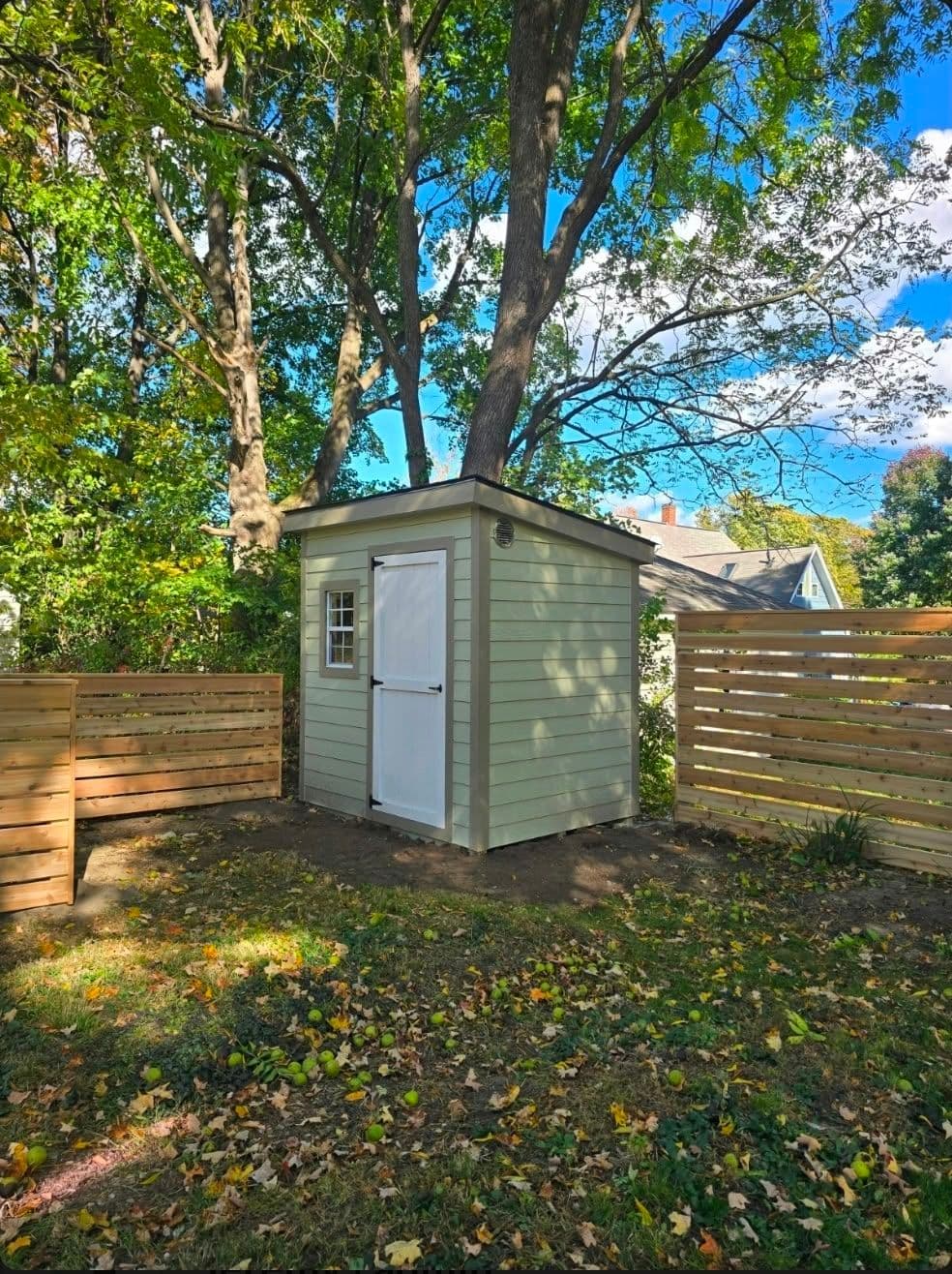 A sage green lap-sided lean-to shed with a white single door and a small window, between two horizontal cedar fences