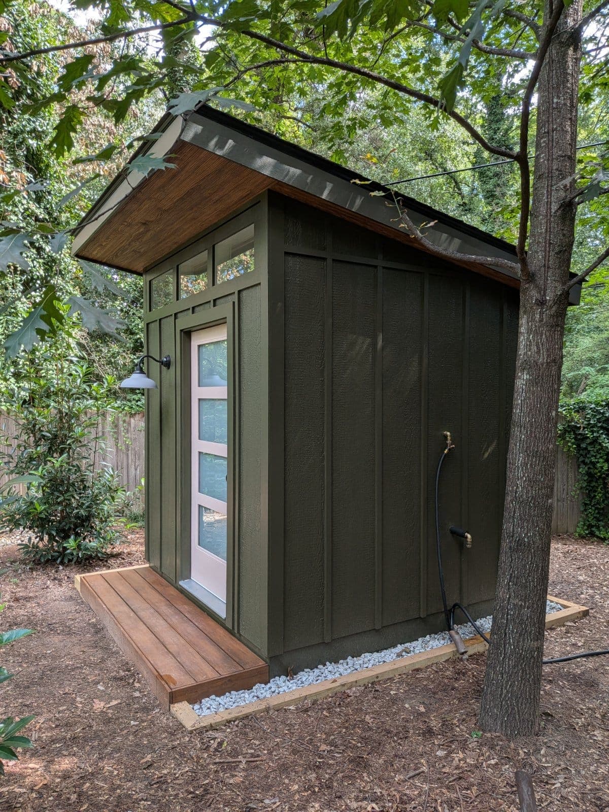 A dark green board-and-batten bathhouse with a frosted-glass door, a wood deck, and a gooseneck light under oak trees
