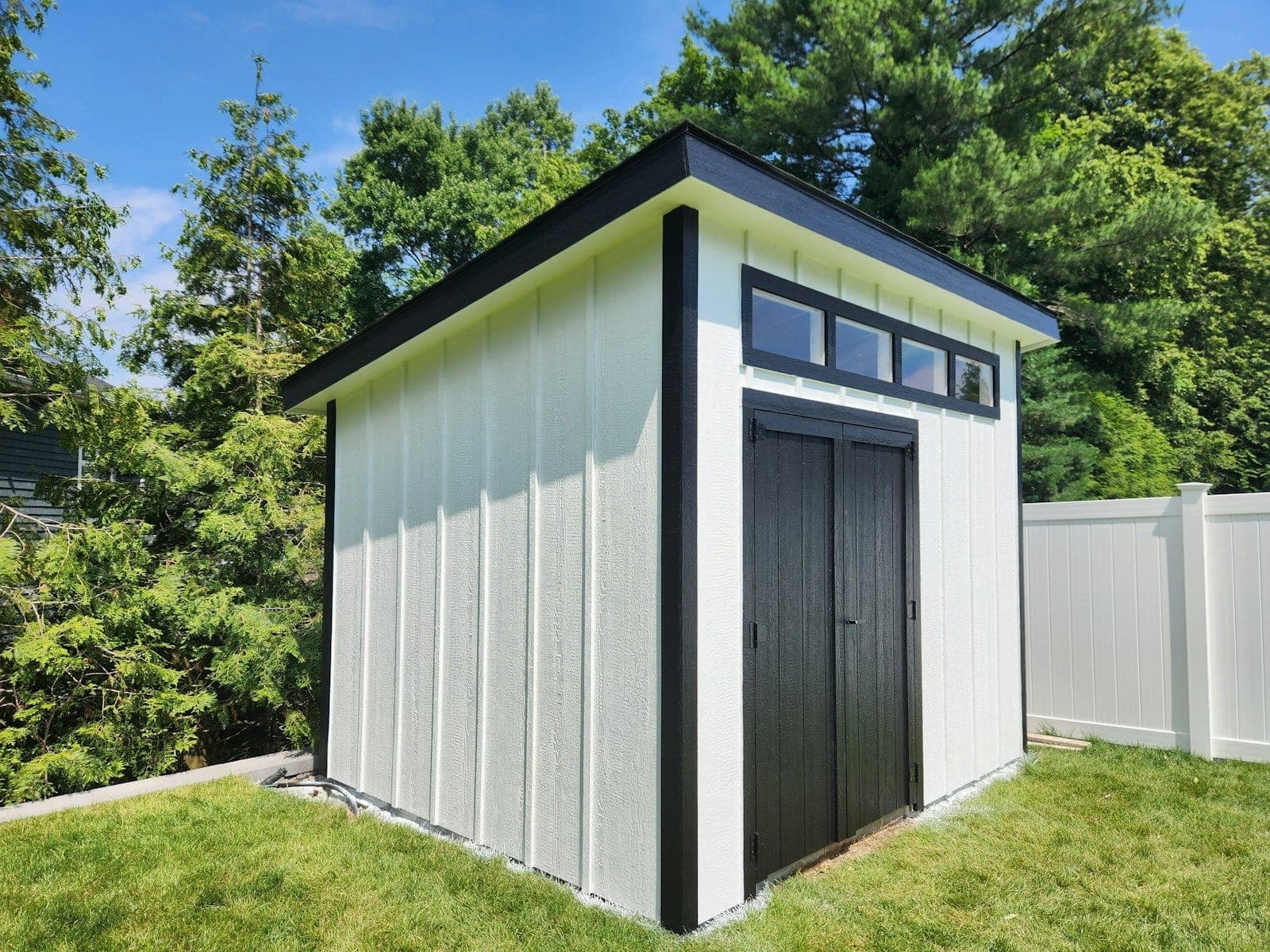 A white board-and-batten shed with black trim and a transom window row, in a fenced suburban backyard